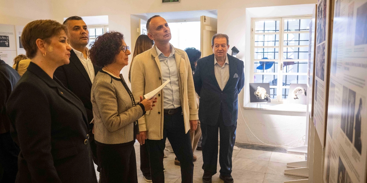 Şapka Devrimi'nin 100. yılına özel düzenlenen sergide, bir grup ziyaretçi duvardaki bilgilendirme panolarını ve sergilenen şapka örneklerini inceliyor. Ön planda, elinde bir metin tutan gözlüklü bir kadın, yanındaki erkeklerle birlikte panodaki görselleri ve metinleri dikkatle takip ediyor. Arka planda, pencere önündeki stantlarda sergilenen farklı şapka modelleri ve tarihi fotoğraflarla dolu sergi panoları yer alıyor.