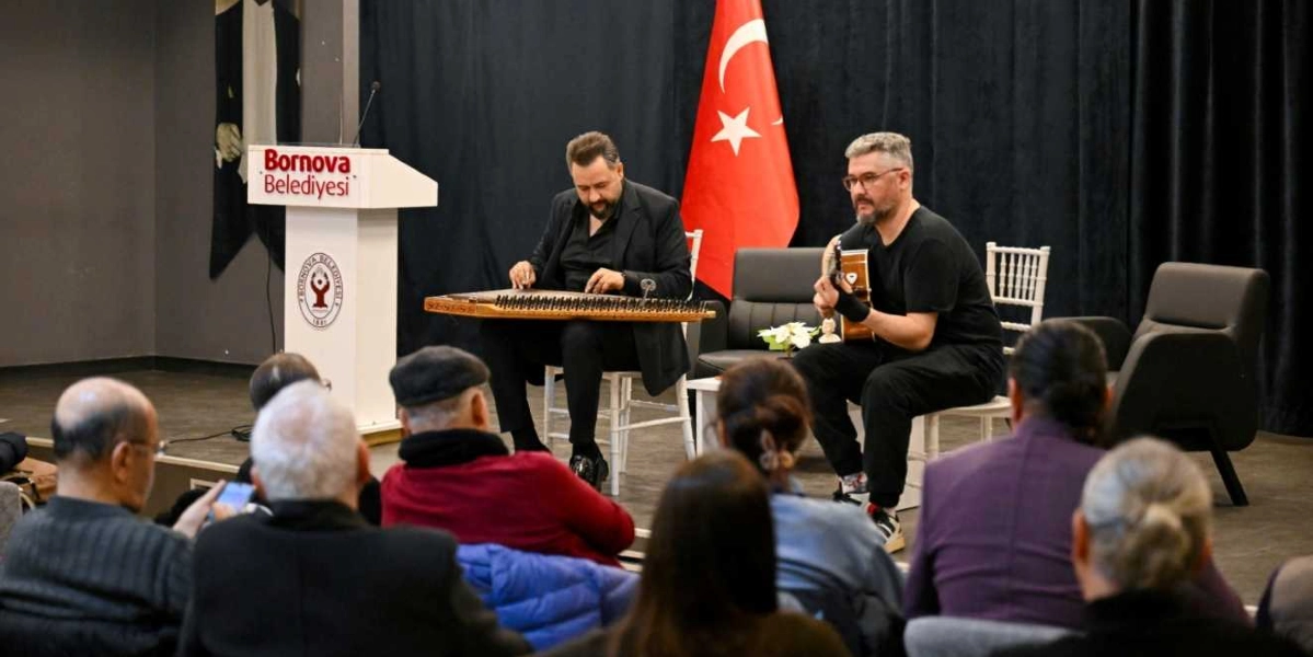 Bornova Belediyesi'ne ait bir sahnede, üzerinde Bornova Belediyesi logosu bulunan beyaz bir kürsünün yanında, biri kanun diğeri gitar çalan iki müzisyen oturuyor. Müzisyenlerin arkasında siyah bir perde ve asılı duran bir Türk bayrağı bulunuyor. İzleyiciler, sahnedeki bu müzik dinletisini dikkatle takip ediyor.