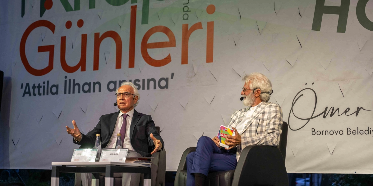 Bornova Belediyesi tarafından düzenlenen etkinlikte, sahnede konuşma yapan iki erkek katılımcı yer alıyor. Solda, koyu renk takım elbiseli ve mor kravatlı, gözlüklü bir konuşmacı ellerini kullanarak bir şeyler anlatıyor. Sağda ise beyaz saçlı ve sakallı, kareli ceketli bir katılımcı elinde renkli bir kitapçıkla onu dinliyor. Arka planda üzerinde 