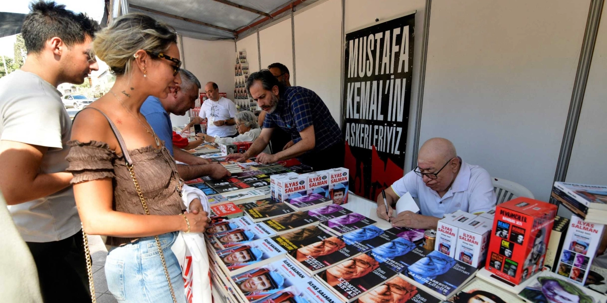 Bir kitap fuarı standında, İlyas Salman masasında oturmuş kitaplarını imzalarken, önündeki stantta İlyas Salman'ın çeşitli kitapları sergileniyor. Standın arka duvarında siyah zemin üzerine beyaz harflerle 
