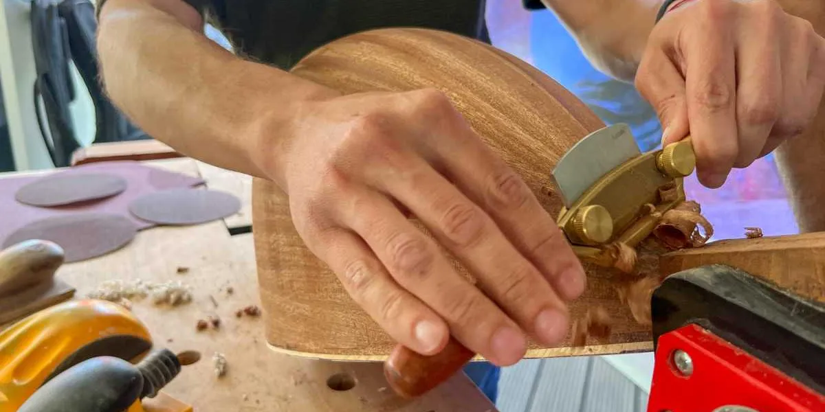 Bir zanaatkar, ahşap bir bağlama teknesini küçük bir el rendesi kullanarak özenle yontuyor. Çalışma masasının üzerinde zımpara kağıtları ve çeşitli el aletleri bulunuyor.