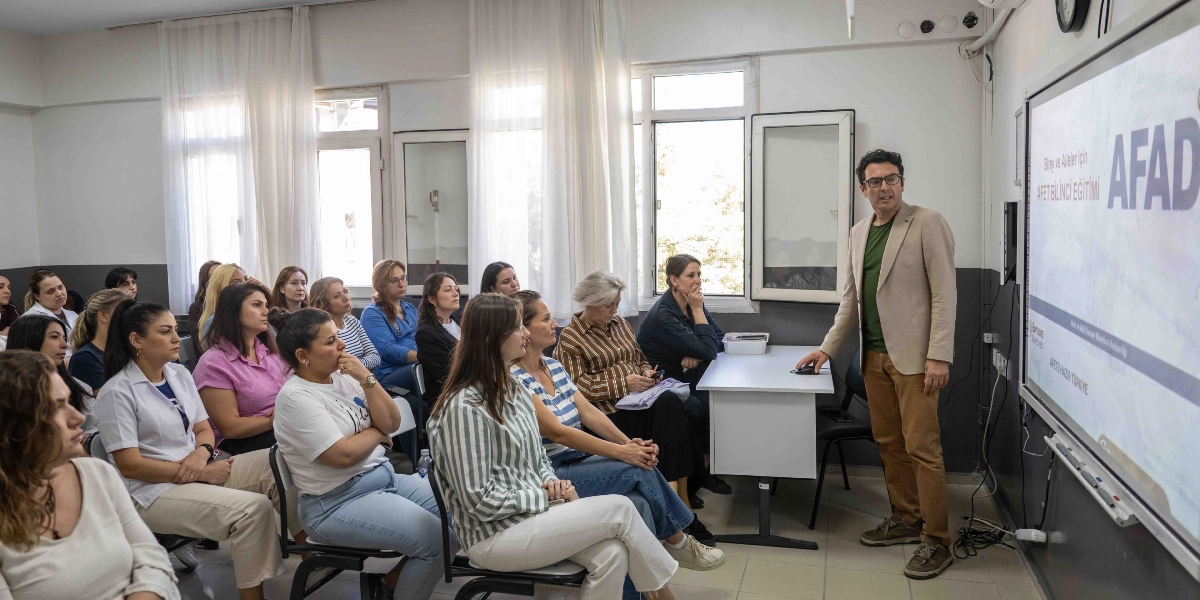 Aydınlık bir sınıfta, gözlüklü bir eğitmen akıllı tahta önünde durarak katılımcılara sunum yapıyor. Akıllı tahtada 