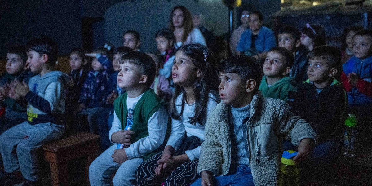 Karanlık bir ortamda, dikkatle ve merakla karşıya bakan bir grup çocuk ve arkalarında oturan yetişkinler. Ön planda, beyaz bluzlu bir kız çocuğu ve yanında bej rengi pelüş ceket giymiş bir erkek çocuğu oturuyor; etraflarında ise farklı yaşlarda birçok çocuk ilgiyle bir gösterimi izliyor.