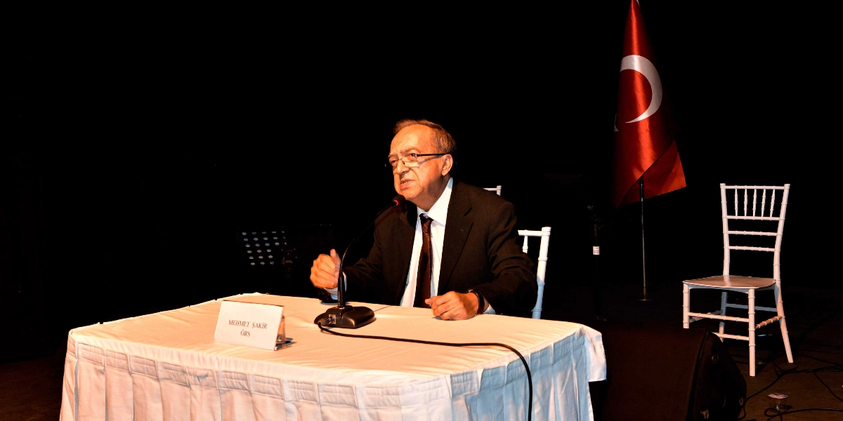 Karanlık bir sahnede, beyaz örtülü bir masanın arkasında oturan ve mikrofonla konuşma yapan gözlüklü, takım elbiseli bir erkek. Masanın üzerinde üzerinde 