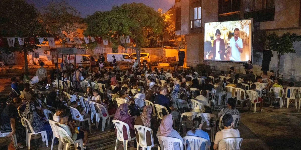 Açık havada, akşam saatlerinde kurulan dev bir ekranda film izleyen kalabalık bir grup insan. İnsanlar beyaz plastik sandalyelerde oturuyor, arka planda ağaçlar ve binalar görülüyor. Ekranın üzerinde Türk bayrakları ve süslemeler asılı.