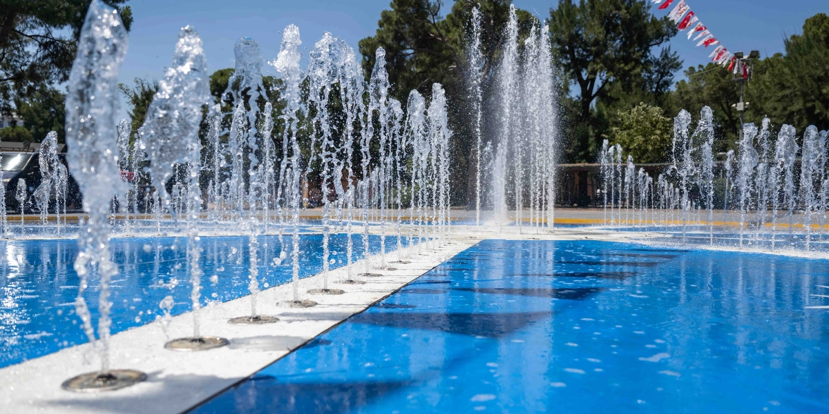 Bornova'da güneşli bir günde, mavi zeminli bir havuzdan yukarı doğru fışkıran çok sayıda su sütunu. Havuzun çevresinde ağaçlar ve havada asılı duran kırmızı beyaz süs flamaları görülüyor.
