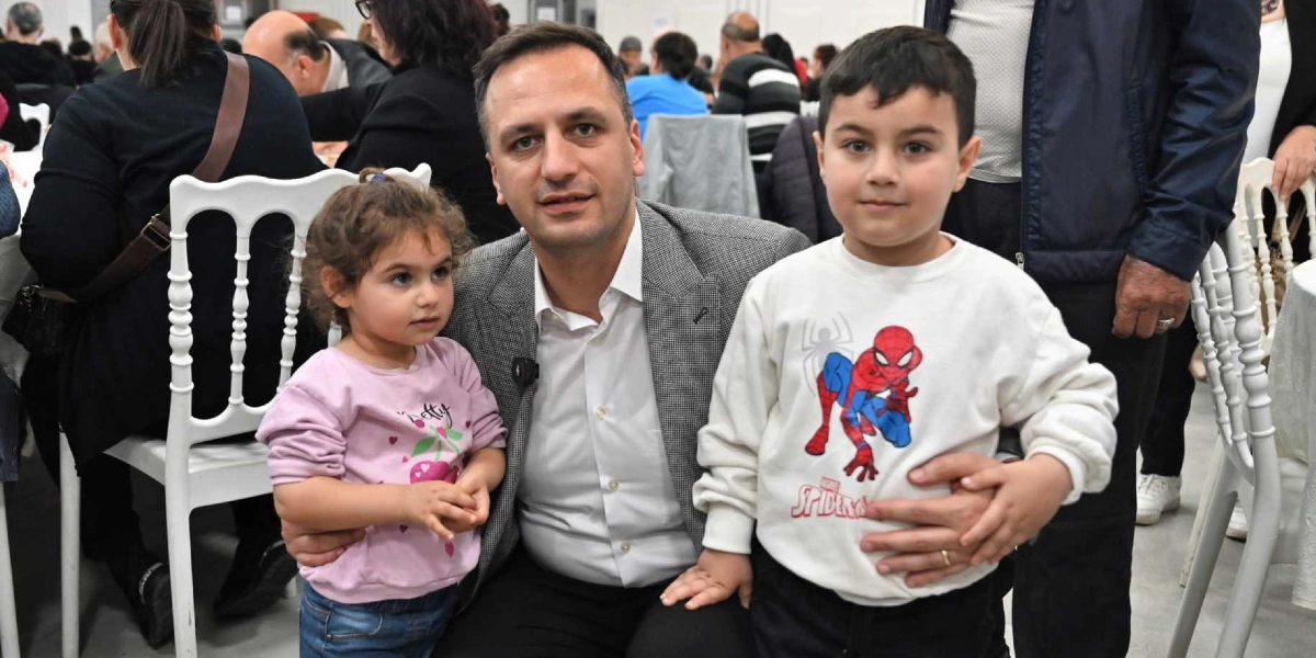 Gri ceketli bir adam, dayanışma yemeğinde yanındaki küçük bir kız çocuğu ve Örümcek Adam baskılı beyaz bir sweatshirt giyen küçük bir erkek çocuğuyla birlikte kameraya gülümseyerek poz veriyor. Arka planda yemek masalarında oturan diğer insanlar ve beyaz sandalyeler görülüyor.