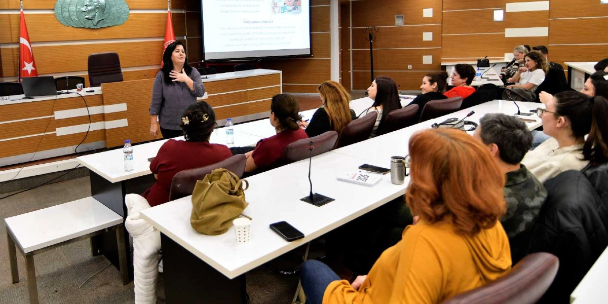 Bornova Belediyesi meclis salonunda, bir konuşmacı ayakta katılımcılara hitap ederken, önündeki uzun masalarda oturan bir grup kadın dikkatle onu dinliyor. Salonun ön tarafındaki projeksiyon perdesinde 