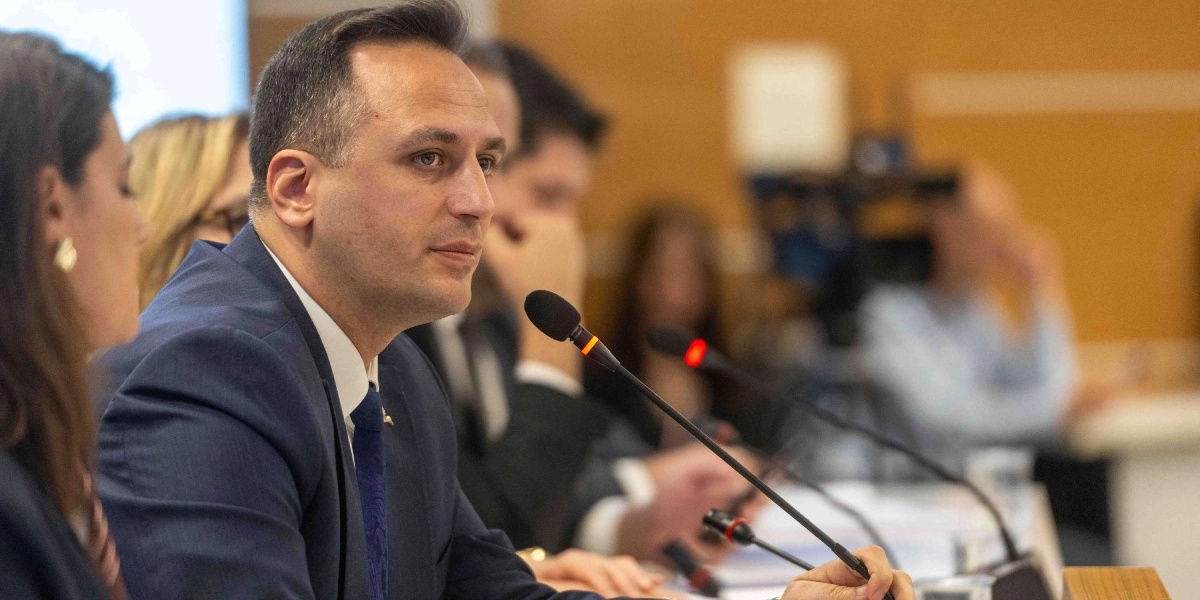 Lacivert takım elbiseli ve mavi kravatlı bir erkek, toplantı masasında önündeki mikrofona konuşuyor. Arka planda bulanık bir şekilde diğer katılımcılar ve toplantı ortamı görünüyor.