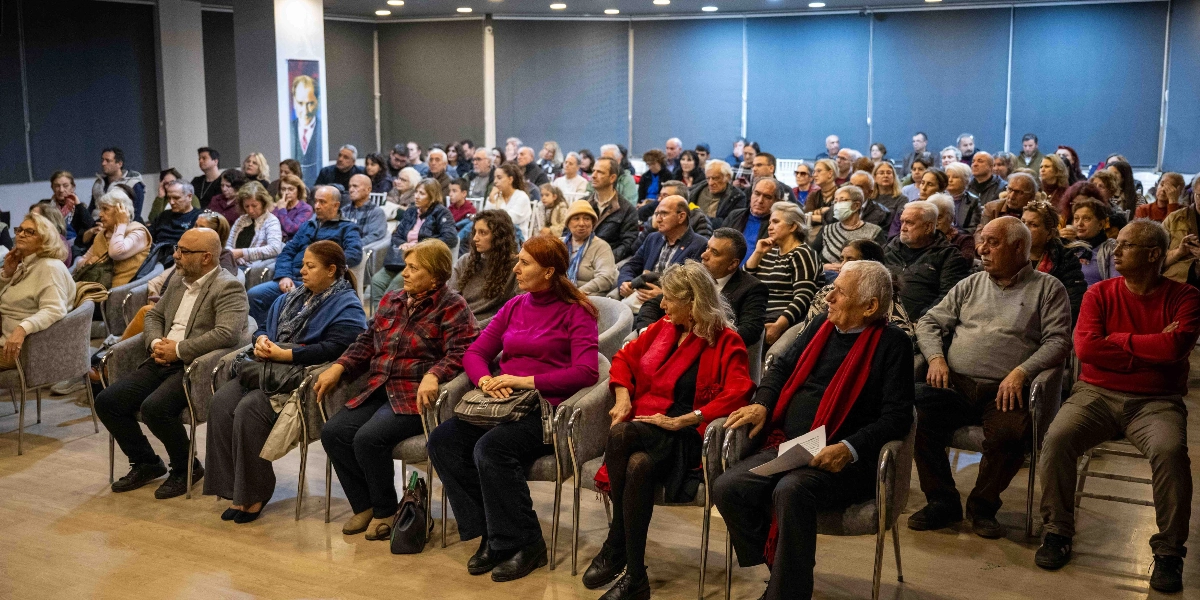Modern bir konferans salonunda, Nazım Hikmet'i anma etkinliği için toplanmış, farklı yaş gruplarından oluşan kalabalık bir dinleyici grubu sandalyelerde oturarak sahneye doğru bakıyor. Salonun arka duvarında Mustafa Kemal Atatürk'ün portresi asılı duruyor. Katılımcıların çoğu günlük kıyafetler içinde, dikkatle dinleme halindeler; ön sıralarda oturan bazı kişiler kırmızı renkli kıyafetler veya aksesuarlar tercih etmiş. Salonun aydınlatması yumuşak ve ortamda ciddi, huzurlu bir atmosfer hakim.