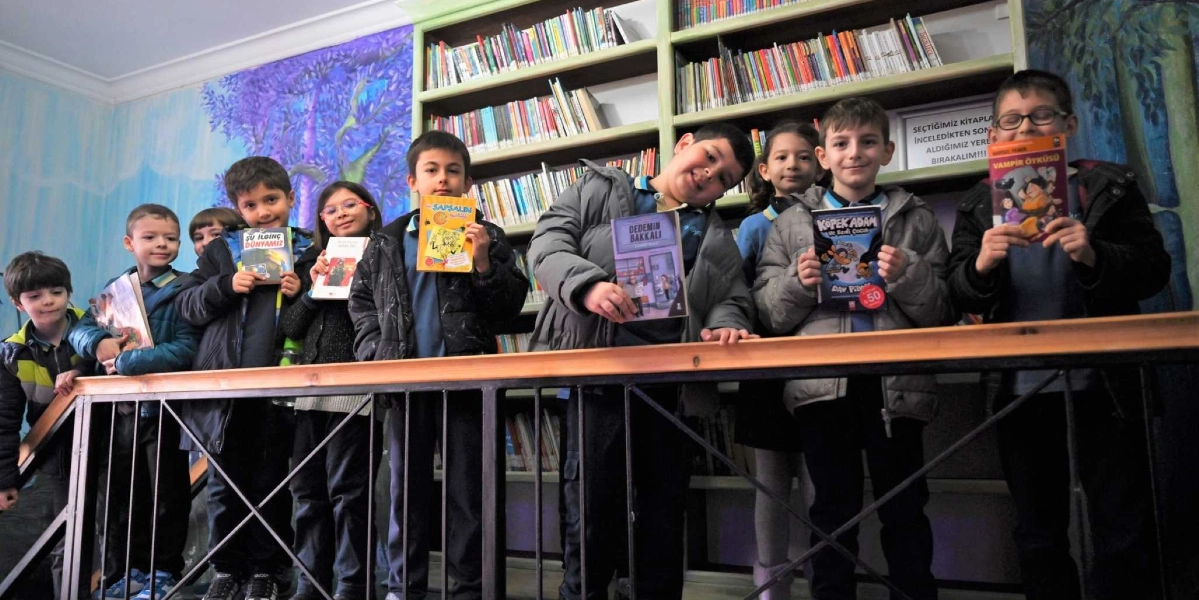 Kütüphanede ellerinde tuttukları çeşitli çocuk kitaplarıyla poz veren bir grup ilkokul öğrencisi. Çocuklar, kitap raflarının önündeki ahşap korkuluklu bir platformda yan yana duruyorlar. Rafların üzerinde 