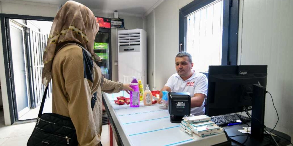 Başörtülü bir kadın, üzerinde Kent Kart logolu beyaz bir tişört bulunan görevliye temizlik ürünleri ve gıda maddelerinden oluşan alışverişini teslim ediyor. Görevli, beyaz bir bankonun arkasında, bilgisayar ekranı ve barkod okuyucunun bulunduğu kasada ödeme işlemini gerçekleştiriyor.