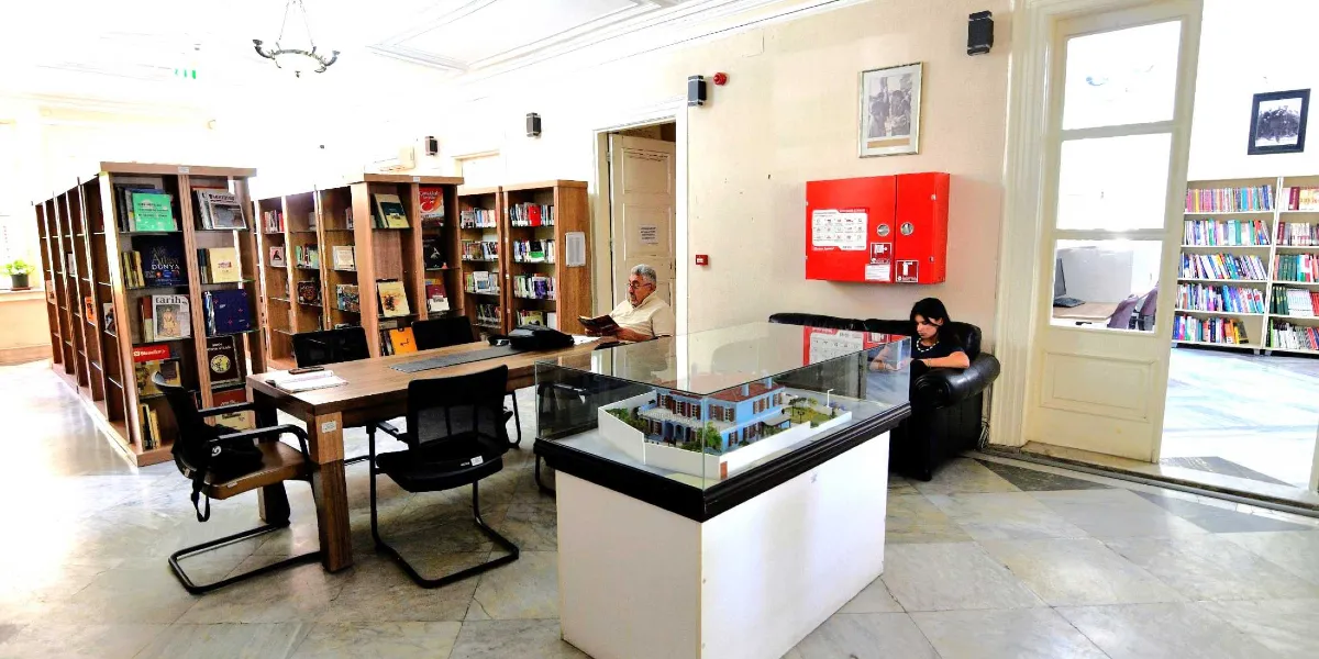 Aydınlık ve yüksek tavanlı bir kütüphane salonunda, ahşap kitaplık rafları, çalışma masaları ve oturma alanları yer alıyor. Salonun ortasında, cam bir fanus içinde tarihi bir köşkün mimari maketi sergileniyor. Arka planda bir kişi masada oturmuş kitap okurken, yan taraftaki siyah deri koltukta oturan bir kadın elindeki telefonla ilgileniyor. Duvarlarda çerçeveli fotoğraflar ve kırmızı bir yangın dolabı bulunuyor. Sağ taraftaki geniş kapıdan, rafları kitaplarla dolu başka bir kütüphane bölümü görünüyor.