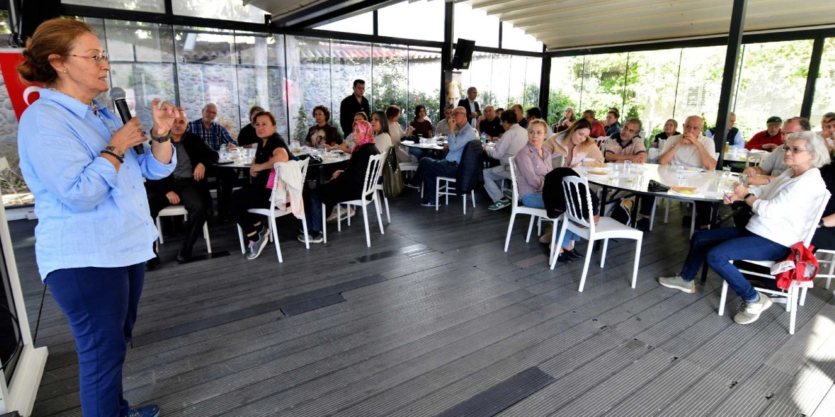 Açık havada, cam panellerle çevrili bir mekanda, mavi gömlekli bir kadın elinde mikrofonla konuşma yaparken, masalarda oturan kalabalık bir grup onu dinliyor.
