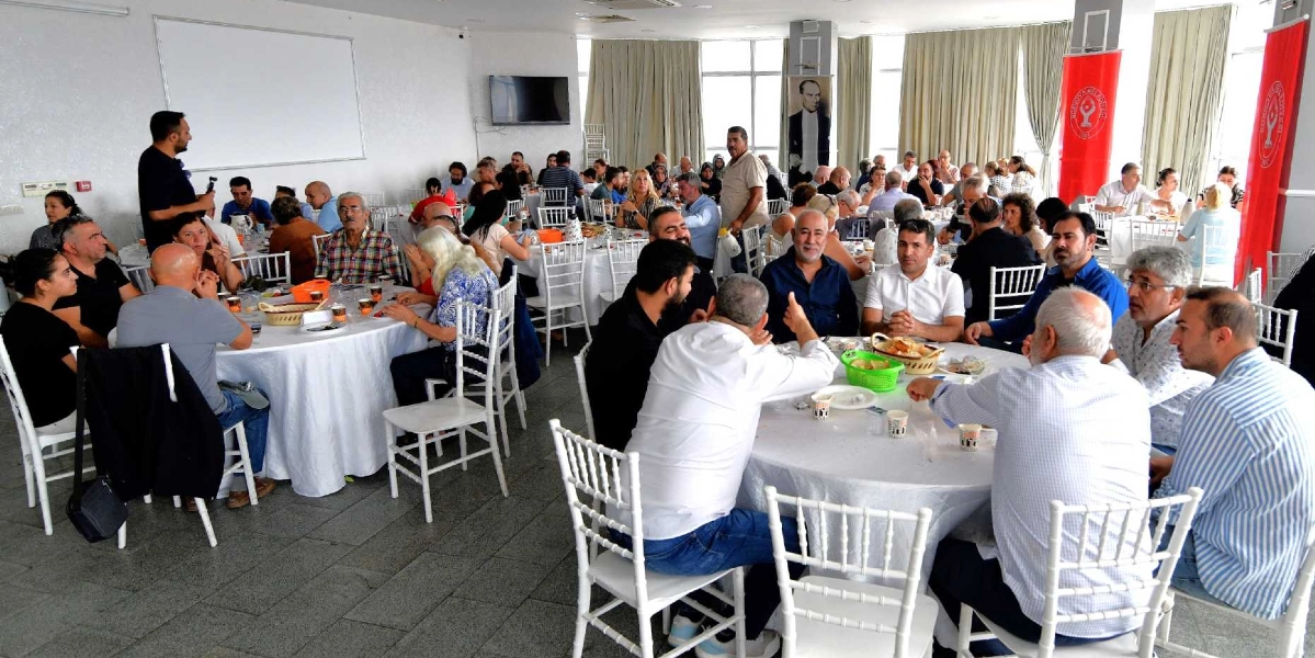 Aydınlık ve geniş bir salonda, beyaz örtülü yuvarlak masalar etrafında toplanmış kalabalık bir grup insan bir arada oturuyor. Salonun ön tarafında ayakta duran bir kişi konuşma yaparken, masalarda oturan katılımcılar onu dinliyor. Duvarlarda Atatürk posteri ve üzerinde Alevi Güçbirliği logosu bulunan kırmızı flamalar asılı. Genel atmosfer, dayanışma ve birliktelik mesajı veren bir toplantı ortamını yansıtıyor.