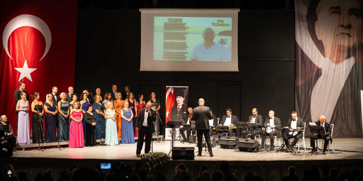 Sahne üzerinde, arkada dev bir Türk bayrağı ve Mustafa Kemal Atatürk'ün portresinin yer aldığı bir konser etkinliği. Sahnenin sol tarafında şık kıyafetler giymiş bir koro, sağ tarafında ise enstrümanlarıyla oturan orkestra üyeleri bulunuyor. Sahnenin ortasında mikrofonuyla şarkı söyleyen bir solist ve ona sırtı dönük şekilde orkestrayı yöneten bir şef yer alıyor. Arka plandaki projeksiyon perdesinde 