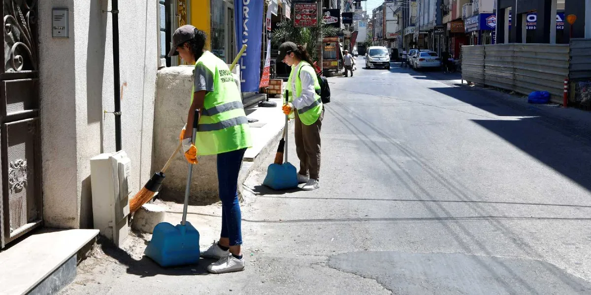 Üzerinde Bornova Belediyesi Temizlik İşleri yazan fosforlu sarı yelekler giymiş iki temizlik görevlisi, bir cadde kenarındaki bina duvarının dibini süpürerek temizlik yapıyor. Görevliler ellerindeki süpürgeler ve mavi faraşlarla çalışırken, arka planda dükkanların bulunduğu hareketli bir şehir caddesi ve araç trafiği görülüyor.