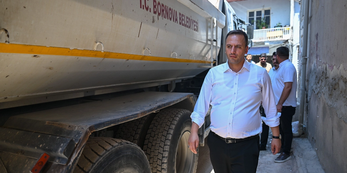 Bornova Belediye Başkanı Ömer Eşki, üzerinde T.C. Bornova Belediyesi yazan bir damperli kamyonun yanında, dar bir sokakta yürürken görülüyor. Başkan beyaz gömlek ve siyah pantolon giymiş, yanında ise beyaz tişörtlü çalışma arkadaşları bulunuyor.