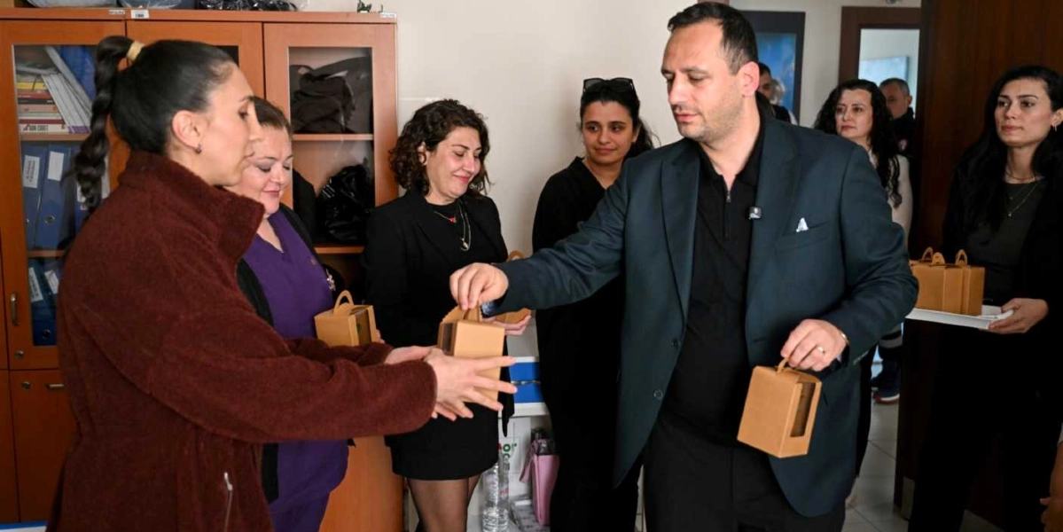 Ofis ortamında, koyu renk ceket giymiş bir erkek, yanında bulunan kadınlara içinde Sağlık Destek Kartı olduğu tahmin edilen küçük, kahverengi karton kutuları dağıtıyor. Arka planda ahşap bir dosya dolabı ve çeşitli giysiler içindeki grup üyeleri yer alıyor.