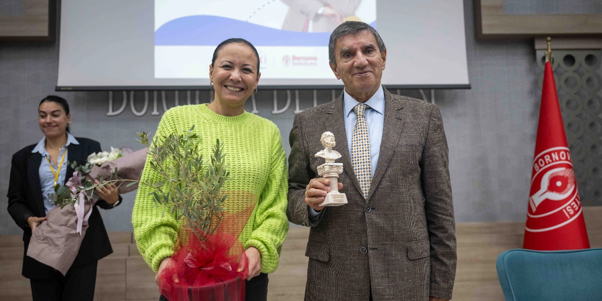 Yeşil kazaklı bir kadın elinde küçük bir zeytin fidanı tutarken, yanında takım elbiseli bir erkek elinde küçük bir büst ile poz veriyor. Arka planda Bornova Belediyesi logosunun bulunduğu bir projeksiyon ekranı ve sağ tarafta Bornova Belediyesi bayrağı yer alıyor. Sol tarafta ise elinde çiçek buketi tutan başka bir kadın gülümsüyor.