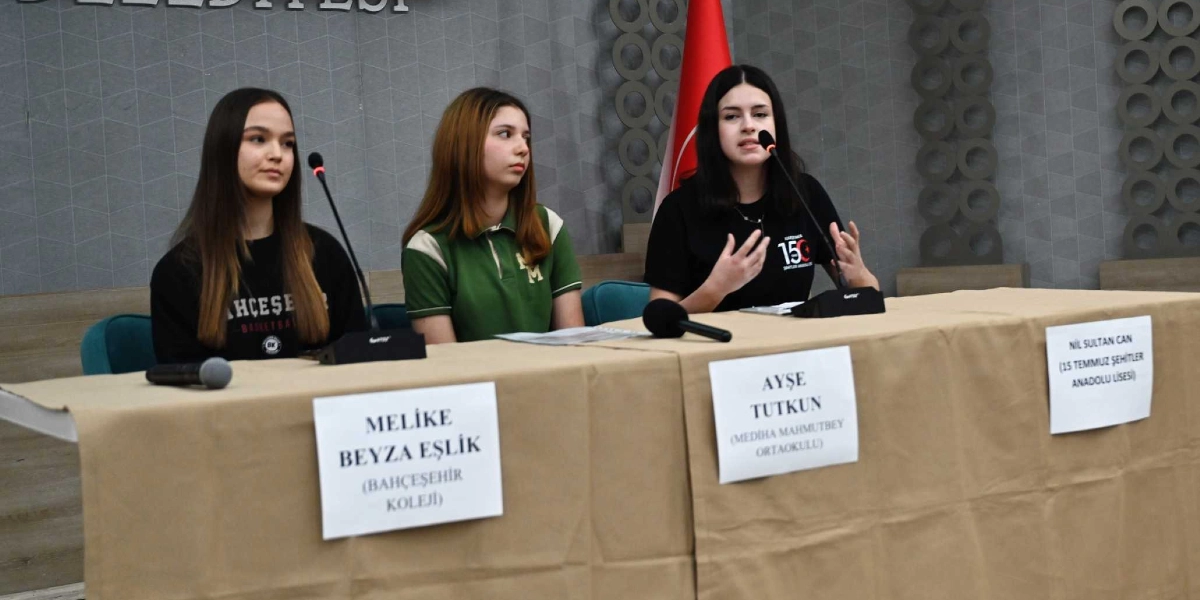 Bej renkli bir masa örtüsüyle kaplı masada yan yana oturan üç genç kız, önlerindeki mikrofonlarla bir konuşma yapıyor. Soldan sağa doğru sırasıyla Bahçeşehir Koleji'nden Melike Beyza Eşlik, Mediha Mahmutbey Ortaokulu'ndan Ayşe Tutkun ve 15 Temmuz Şehitler Anadolu Lisesi'nden Nil Sultan Can yer alıyor. Masanın ön kısmında her birinin isminin ve okulunun yazılı olduğu beyaz kağıtlar bulunuyor. Arka planda gri duvar üzerinde kısmen görünen belediye yazısı ve Türk bayrağı yer alıyor.