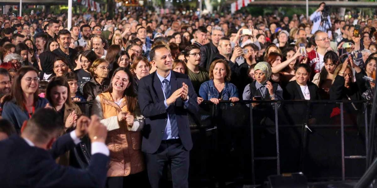 Bornova'da düzenlenen Gençlik Haftası etkinlikleri kapsamında, sahne önündeki bariyerlerin arkasında toplanmış kalabalık bir izleyici grubu. Ön planda, lacivert takım elbiseli ve kravatlı bir erkek alkışlayarak sahneyi izliyor; yanında ise gülümseyen ve yine alkışlayan kadınlar ile diğer izleyiciler yer alıyor. Arka planda ise oldukça yoğun, sahne performansını izleyen ve bazıları telefonlarıyla kayıt yapan çok sayıda genç ve yetişkin bulunuyor.