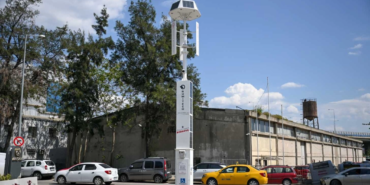 Bornova'da bir cadde kenarında, üzerinde güneş panelleri ve dikey rüzgar türbini bulunan, Rumitech markalı hibrit bir enerji üretim direği yer alıyor. Direğin gövdesinde Bornova Belediyesi logosu ve 