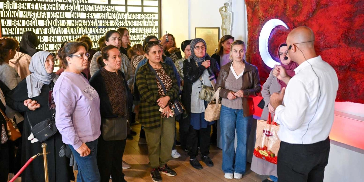 Belkahve'deki bir müze veya sergi alanında, beyaz gömlekli ve mikrofonlu bir rehberi dikkatle dinleyen bir grup kadın. Arka planda duvarda Mustafa Kemal Atatürk'ün sözlerinin yer aldığı büyük bir pano ve sağ tarafta ışıklandırılmış kırmızı zemin üzerine beyaz ay-yıldız figürü bulunuyor. Katılımcılar günlük kıyafetleriyle, rehberin anlatımını takip ederek sergiyi geziyor.