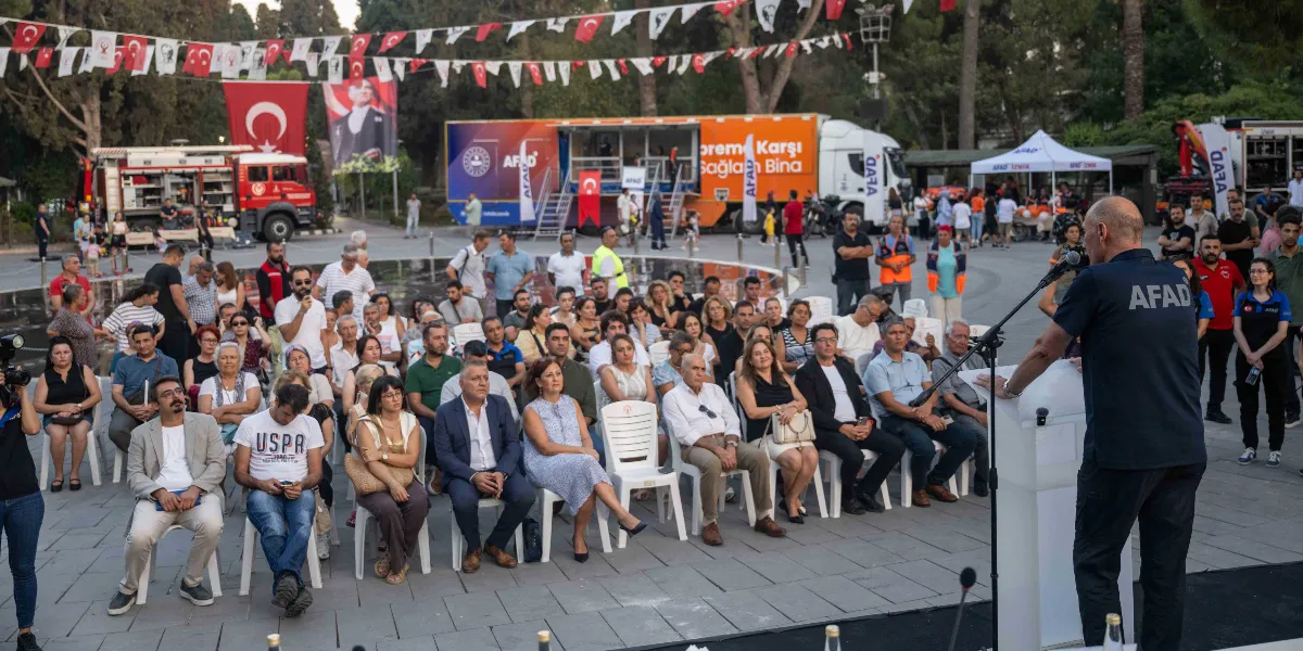 Açık havada düzenlenen bir afet bilinci toplantısında, üzerinde AFAD yazılı tişört giyen bir konuşmacı kürsüde hitap ederken, önünde oturan kalabalık bir grup kendisini dinliyor. Arka planda üzerinde 
