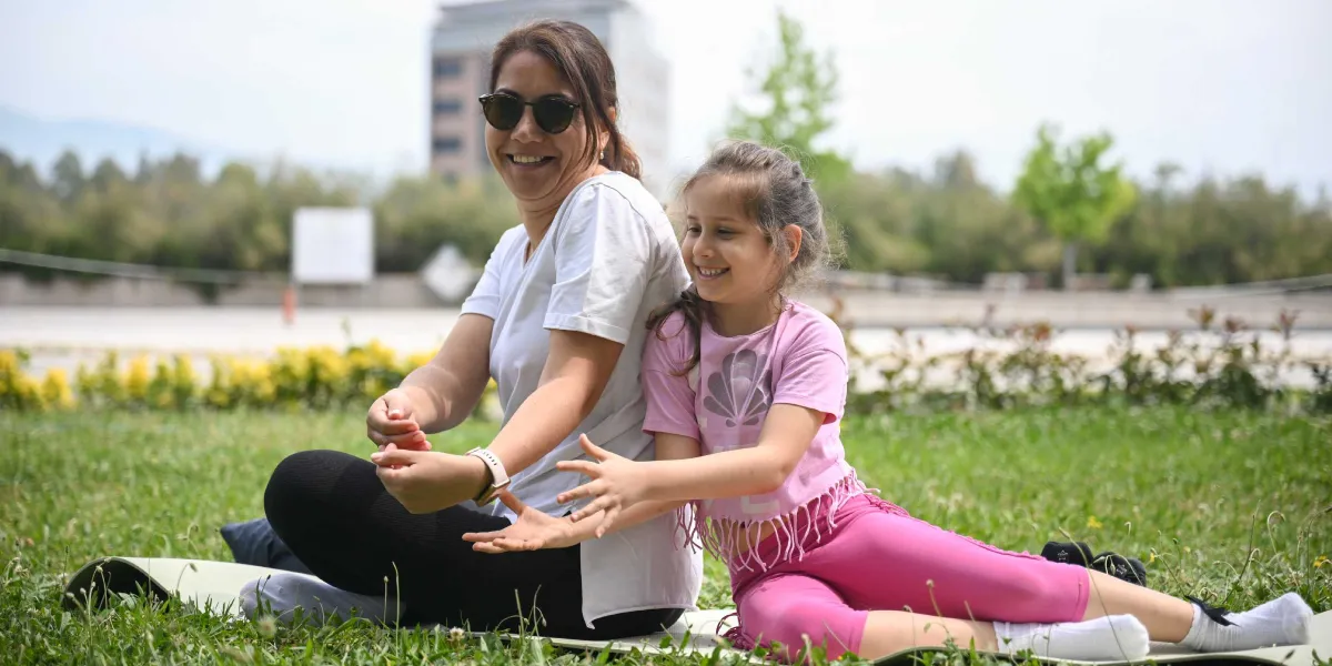 Açık havada, yeşil bir çim alan üzerinde sırt sırta oturmuş, gülümseyen bir anne ve kızı. Beyaz tişörtlü ve güneş gözlüklü anne ile pembe tişörtlü ve pembe taytlı küçük kız çocuğu, elleriyle bir oyun oynar gibi birbirlerine doğru uzanmış neşeli bir şekilde vakit geçiriyor.