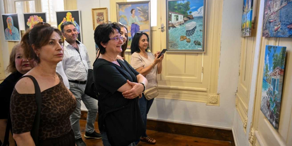 Bir sanat galerisinde duvarlara asılı tabloları ilgiyle inceleyen bir grup insan. Ön planda, leopar desenli bir bluz giyen kadın ve yanında siyah tişörtlü, gözlüklü bir kadın yer alıyor. Arkalarında ise beyaz gömlekli bir erkek ve telefonla fotoğraf çeken başka bir kadın bulunuyor. Duvarlarda deniz manzaralı, figüratif ve renkli yağlı boya tablolar sergileniyor.