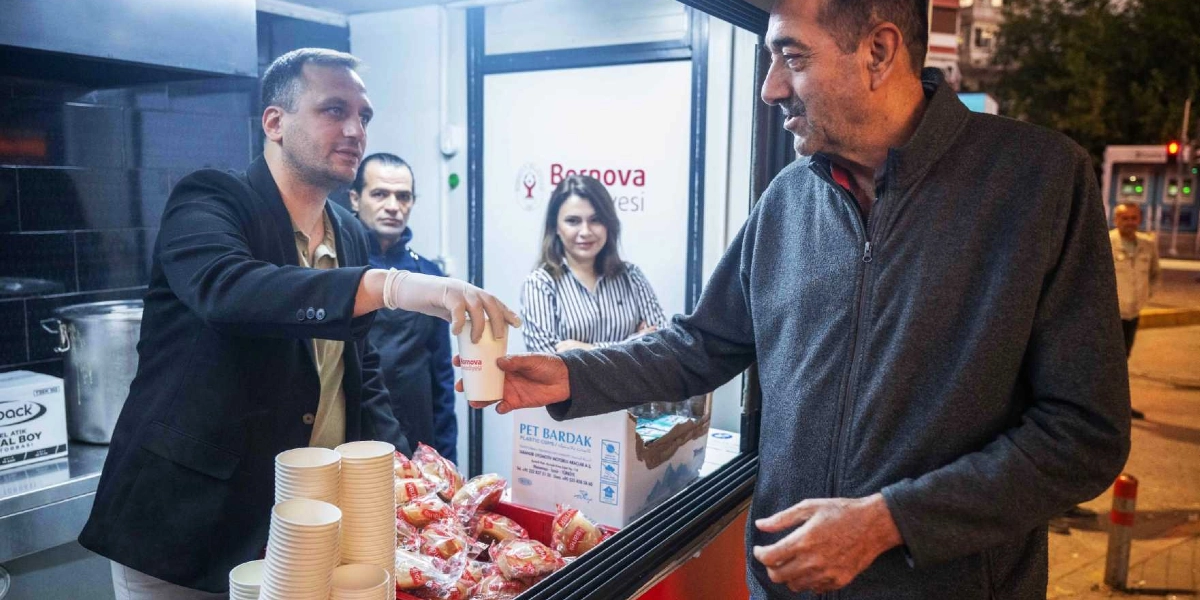 Bornova Belediyesi tarafından kurulan bir stantta, görevli bir kişi sabah saatlerinde bir vatandaşa üzerinde Bornova Belediyesi logosu bulunan bir karton bardakla sıcak çorba ikram ediyor. Arka planda başka görevlilerin de bulunduğu stantta, yanında paketli ekmeklerin ve üst üste dizilmiş boş karton bardakların olduğu görülüyor.