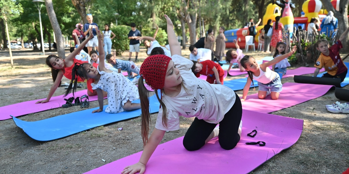 Büyükpark'ta açık havada yoga matları üzerinde egzersiz yapan bir grup çocuk; ön planda kırmızı örgülü şapkalı, beyaz tişörtlü ve siyah taytlı bir kız çocuğu yoga hareketi yaparken, arkasında diğer çocuklar da benzer pozisyonlarda onlara eşlik ediyor, arka planda ise şişme oyun parkı ve ağaçlar görülüyor.