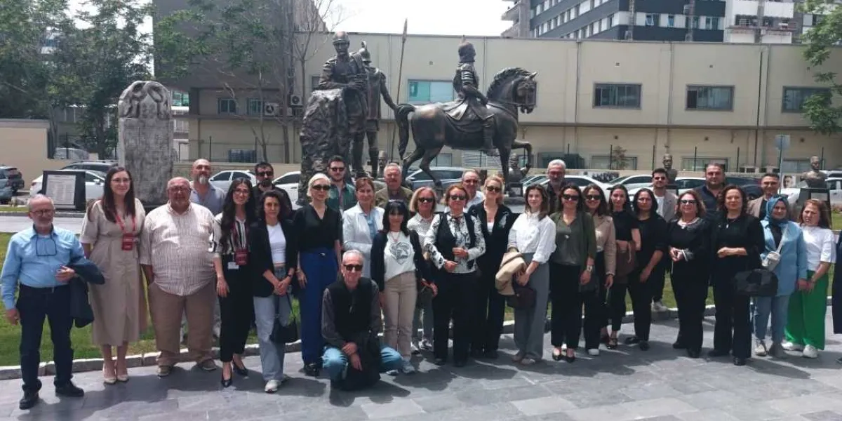 Bornova'da bir açık hava müzesi veya tarihi bir anıtın önünde toplanmış, çeşitli yaş gruplarından oluşan kalabalık bir grup insan, kameraya gülümseyerek poz veriyor. Grubun arkasında, at üzerinde bir şövalyeyi ve yanında duran askerleri tasvir eden bronz bir heykel grubu ile taş bir dikilitaş yer alıyor. Arka planda ise park halindeki araçlar ve modern bir bina yapısı görülüyor.