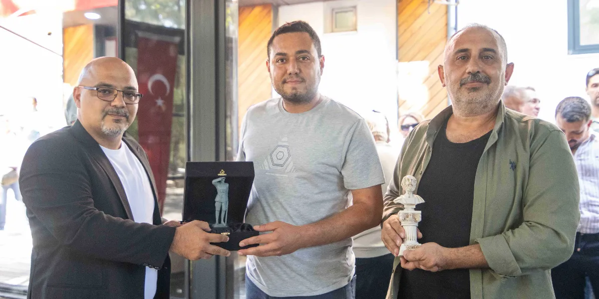 Üç erkek yan yana duruyor. Soldaki gözlüklü ve siyah ceketli kişi, ortadaki gri tişörtlü kişiye içerisinde asker figürü bulunan siyah bir kutu hediye ediyor. Sağdaki kişi ise elinde küçük bir büst tutuyor. Arka planda Türk bayrağı ve bulanık bir kalabalık görülüyor.