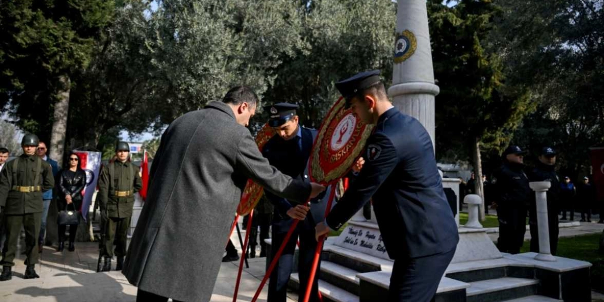 Gri paltolu bir erkek ve iki polis memuru, Bornova Belediyesi yazılı kırmızı çelenkleri mermer bir anıtın önüne yerleştiriyor. Tören alanında askerler, polisler ve sivil katılımcılar hazır bulunuyor; arka planda ağaçlar ve Türk bayrakları yer alıyor.