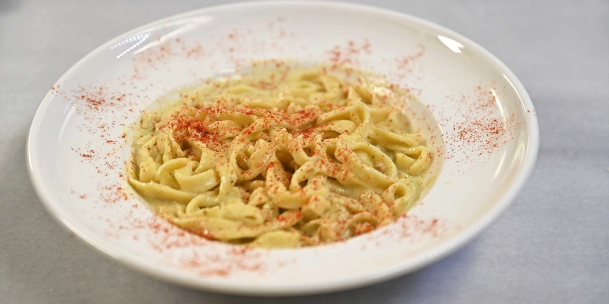 Beyaz, geniş kenarlı bir tabak içerisinde servis edilen, kremsi soslu ve üzerine kırmızı toz baharat serpilmiş fettuccine makarna.