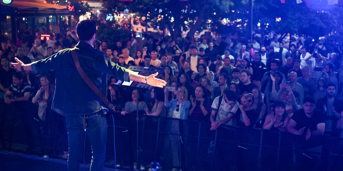 Açık havada düzenlenen bir konserde, sahnedeki erkek sanatçı arkası dönük bir şekilde kollarını iki yana açarak kalabalığa sesleniyor. Mavi ışıklarla aydınlatılan sahnede sanatçının önünde bir nota sehpası bulunuyor. Bariyerlerin arkasında toplanan kalabalık, elleriyle alkış tutarak ve telefonlarıyla çekim yaparak konseri izliyor.