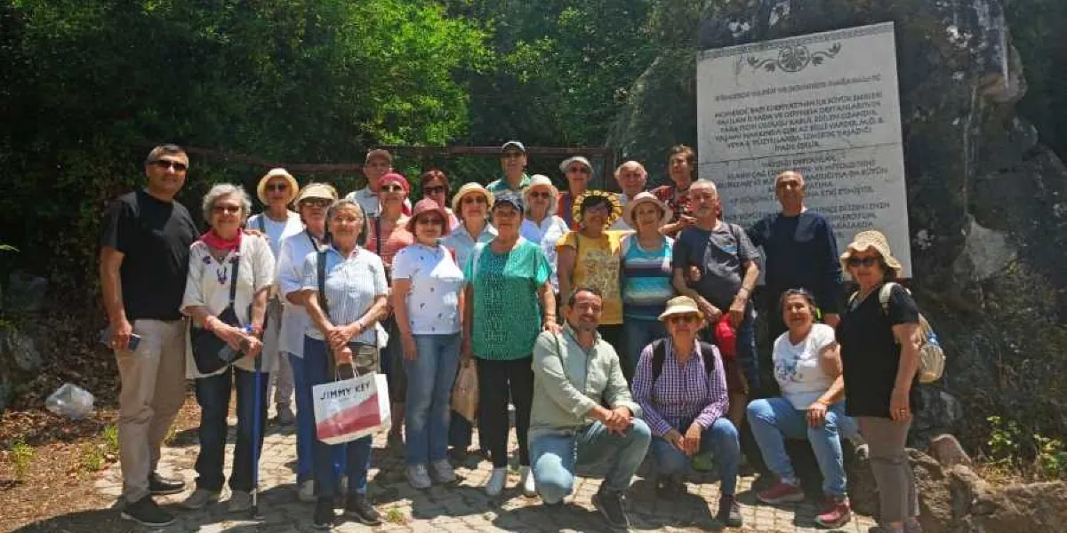 Ağaçlık bir alanda, üzerinde bilgilendirici bir metin bulunan büyük bir taş anıtın önünde yan yana duran, farklı yaş gruplarından oluşan yaklaşık yirmi kişilik bir grup insan. Katılımcıların çoğu günlük kıyafetler ve şapkalar giymiş, bazıları gülümseyerek kameraya bakıyor. Grubun önünde çömelmiş bir erkek, arkasında ise ayakta duran kadın ve erkekler yer alıyor. Anıtın üzerindeki metin, bölgenin tarihi ve doğası hakkında bilgi veriyor.