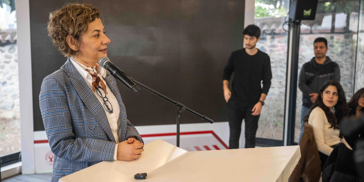 Kıvırcık saçlı, ekose ceketli bir kadın, kürsüde mikrofon başında konuşma yapıyor. Arka planda ayakta duran iki genç erkek ve oturarak onu dinleyen genç kadınlar bulunuyor. Ortam, ÇYDD Bornova şubesindeki bir toplantı salonunu yansıtıyor.