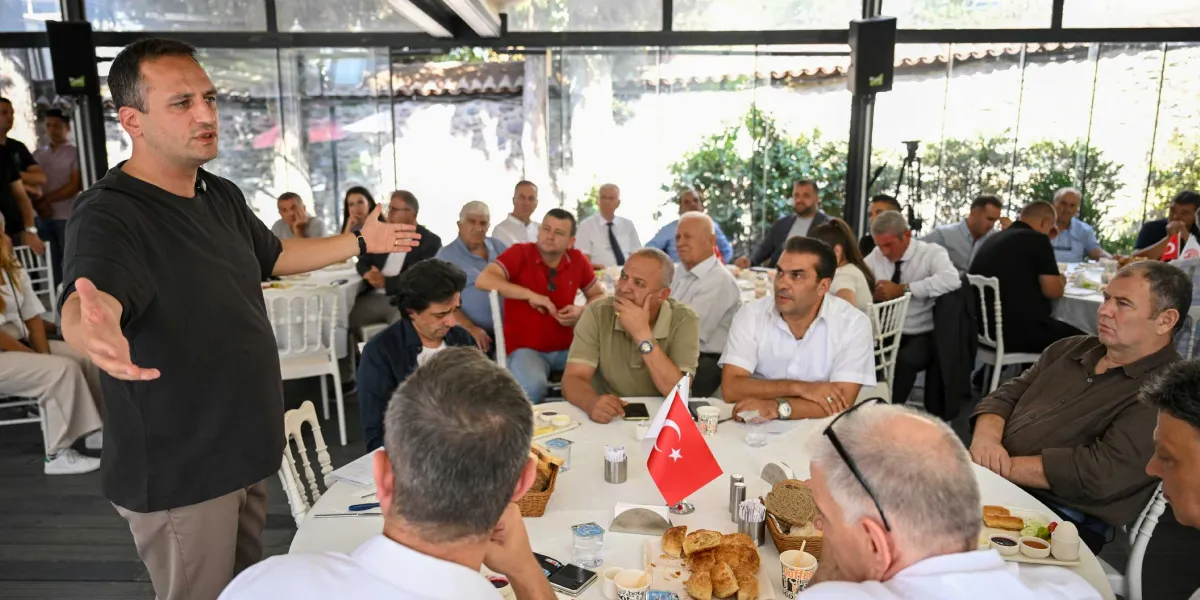 Dramalılar Köşkü’nün camla kaplı, aydınlık bir salonunda düzenlenen toplantıda, siyah tişörtlü bir konuşmacı ayakta durarak elleriyle açıklama yapıyor. Yuvarlak masanın etrafında oturan, aralarında Bornova Belediyesi yetkilileri ve sanayicilerin bulunduğu kalabalık bir grup, konuşmacıyı dikkatle dinliyor. Masaların üzerinde Türk bayrakları, kahvaltılıklar ve içecekler yer alıyor.