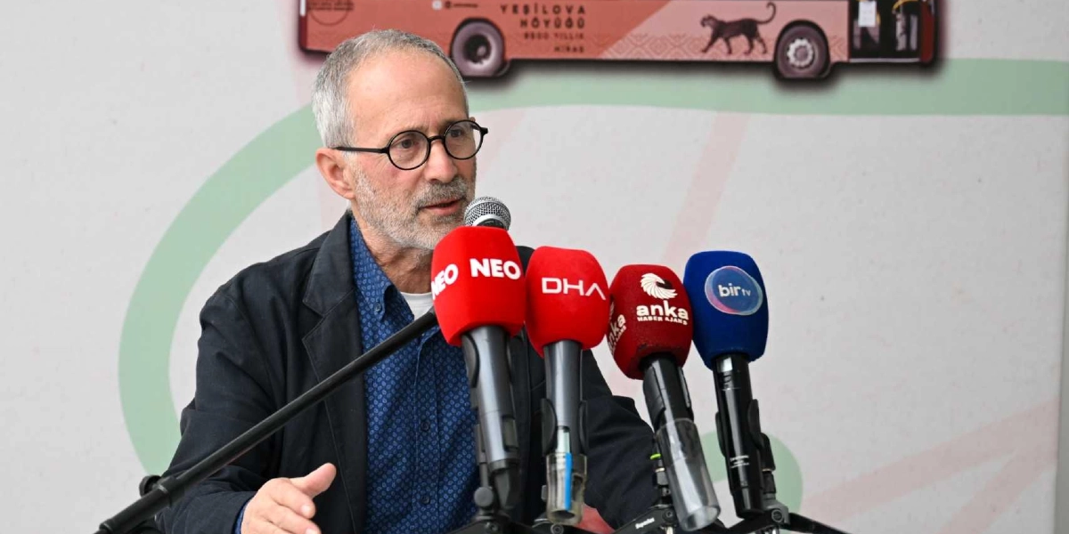 Gözlüklü, kır saçlı ve sakallı bir erkek, önünde Neo, DHA, Anka ve Bir TV mikrofonlarının bulunduğu bir basın toplantısında konuşma yapıyor. Arka planda üzerinde 