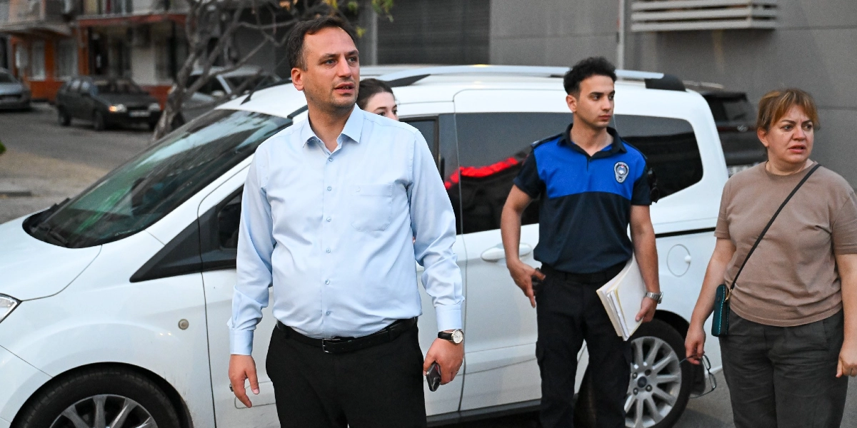 Açık mavi gömlekli bir erkek, yanında üniformalı bir zabıta memuru ve bej tişörtlü bir kadınla birlikte sokakta yürürken görülüyor. Zabıta memurunun elinde bir dosya bulunuyor ve arka planda beyaz bir araç yer alıyor.