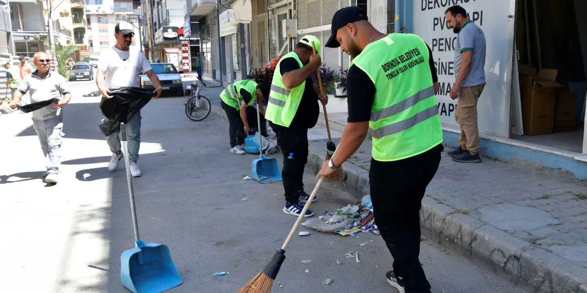 Bornova Belediyesi Temizlik İşleri Müdürlüğü yelekleri giymiş bir grup görevli, sokakta süpürge ve faraşlarla temizlik çalışması yapıyor. Ön planda, üzerinde Bornova Belediyesi Temizlik İşleri Müdürlüğü yazılı neon yeşil yelekli bir görevli sokağı süpürürken, diğer görevliler de çevredeki çöpleri topluyor.