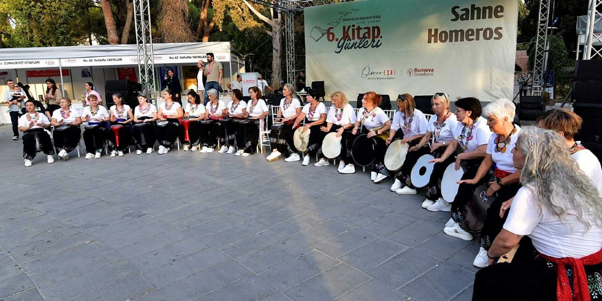 Bornova Belediyesi tarafından düzenlenen 6. Kitap Günleri etkinliğinde, beyaz tişörtlü ve renkli kolyeli kadınlardan oluşan bir ritim grubu, açık havada yan yana dizilmiş sandalyelerde oturarak ellerindeki vurmalı çalgıları çalıyor. Arka planda 