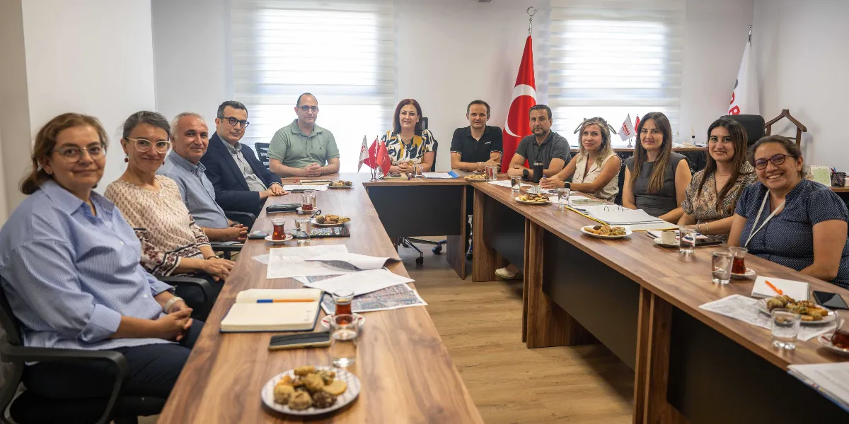 U-şeklinde birleştirilmiş toplantı masasının etrafında oturan, aralarında Bornova Belediyesi yetkililerinin de bulunduğu on bir kişilik grup, toplantı halindeyken kameraya gülümsüyor. Masanın üzerinde çay bardakları, atıştırmalık tabakları, not defterleri ve çeşitli belgeler yer alıyor. Arka planda Türk bayrağı ve Bornova Belediyesi'ne ait olduğu anlaşılan bir flama bulunuyor.