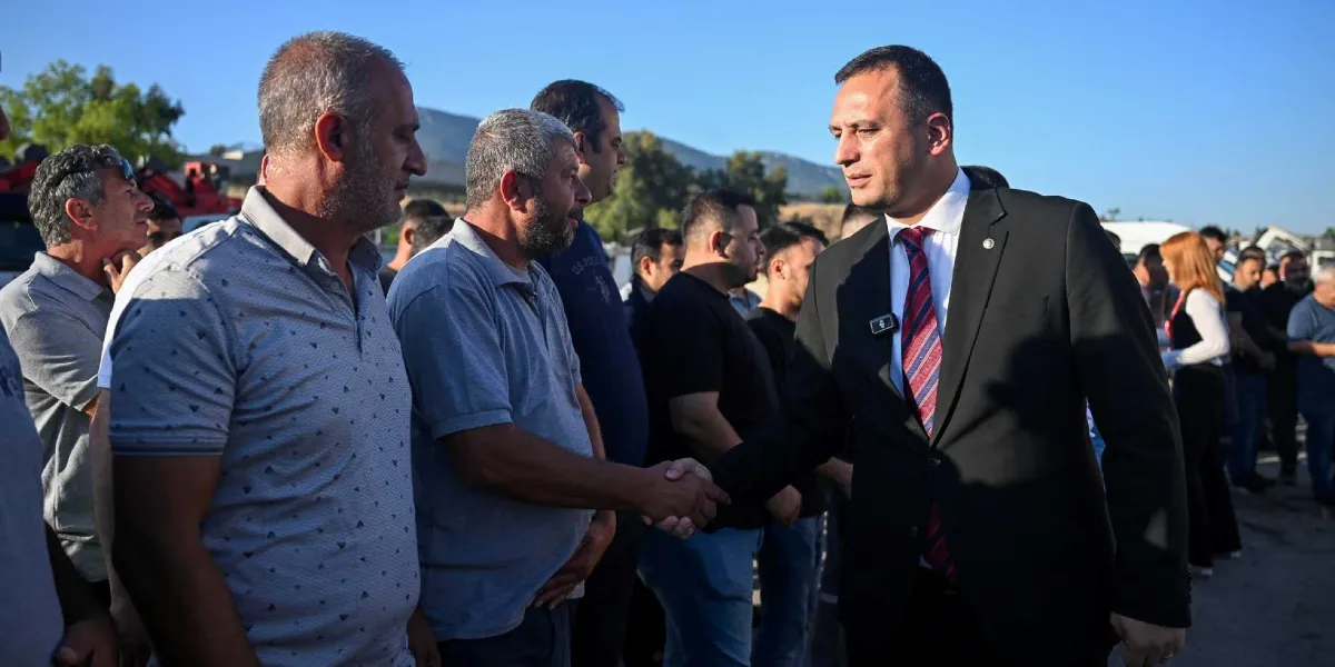 Siyah takım elbiseli ve kırmızı çizgili kravatlı bir erkek, açık havada bir grup erkekle tokalaşıyor. Tokalaştığı kişiler günlük kıyafetler içinde, daha çok işçi veya sendika temsilcisi görünümünde. Arka planda tepeler ve açık bir gökyüzü bulunuyor.