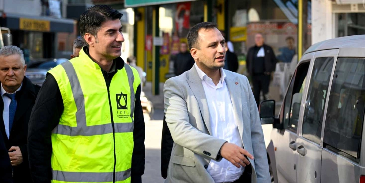 Üzerinde İZSU logosu bulunan fosforlu sarı yelekli bir yetkili ile yanında gri ceketli bir başka yetkili, bir sokakta incelemelerde bulunuyor. Gri ceketli kişi elini ileriye doğru uzatarak bir şeyler anlatırken, yanındaki fosforlu yelekli kişi onu dinliyor. Arka planda ise iş yerleri ve park halindeki araçlar görülüyor.