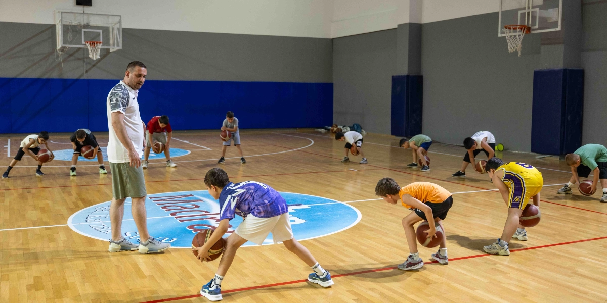 Kapalı bir spor salonunda, antrenör eşliğinde basketbol antrenmanı yapan bir grup çocuk. Antrenör ayakta durarak öğrencileri izlerken, çocuklar parke zemin üzerinde eğilmiş vaziyette ellerinde basketbol toplarıyla top sürme çalışması yapıyor.