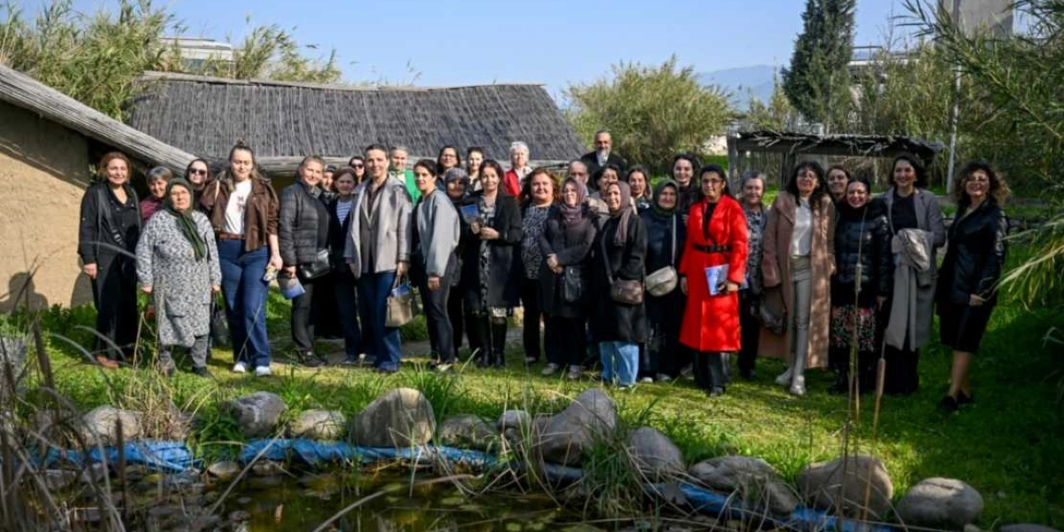 Yeşil bir bahçede, arkalarında sazdan çatılı geleneksel bir yapı bulunan, çoğunluğu kadınlardan oluşan kalabalık bir grup, ön plandaki küçük bir süs havuzunun kenarında yan yana durarak poz veriyor. Gruptaki kişiler günlük kıyafetler ve ceketler giymiş durumda; aralarında kırmızı kabanlı bir kadın dikkat çekiyor. Gün ışığında çekilen bu karede, katılımcılar samimi bir atmosferde bir arada görünüyor.